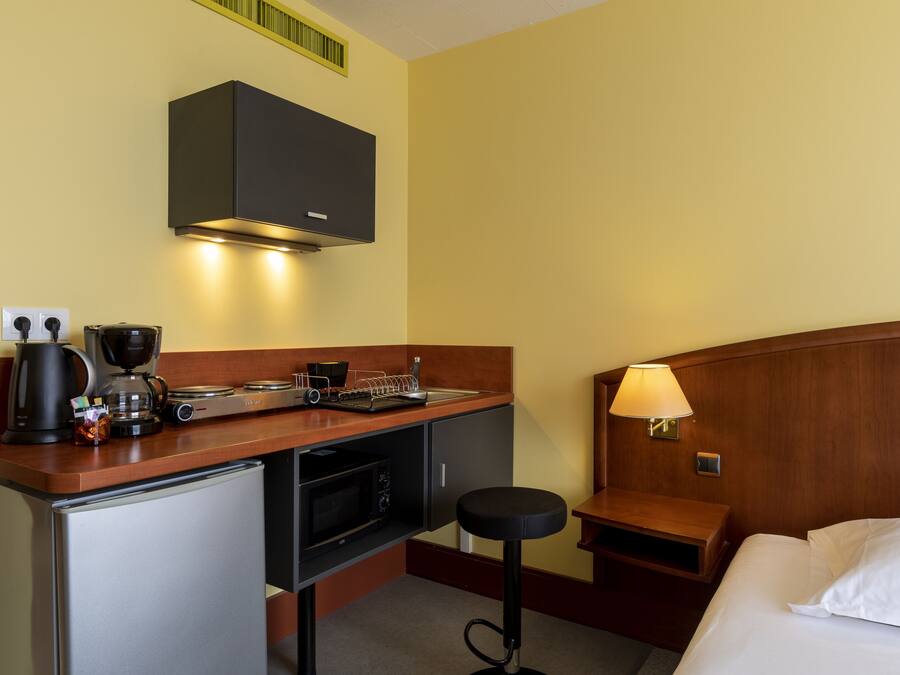 A compact hotel room with a kitchenette, a bed, a nightstand, and a small stool.