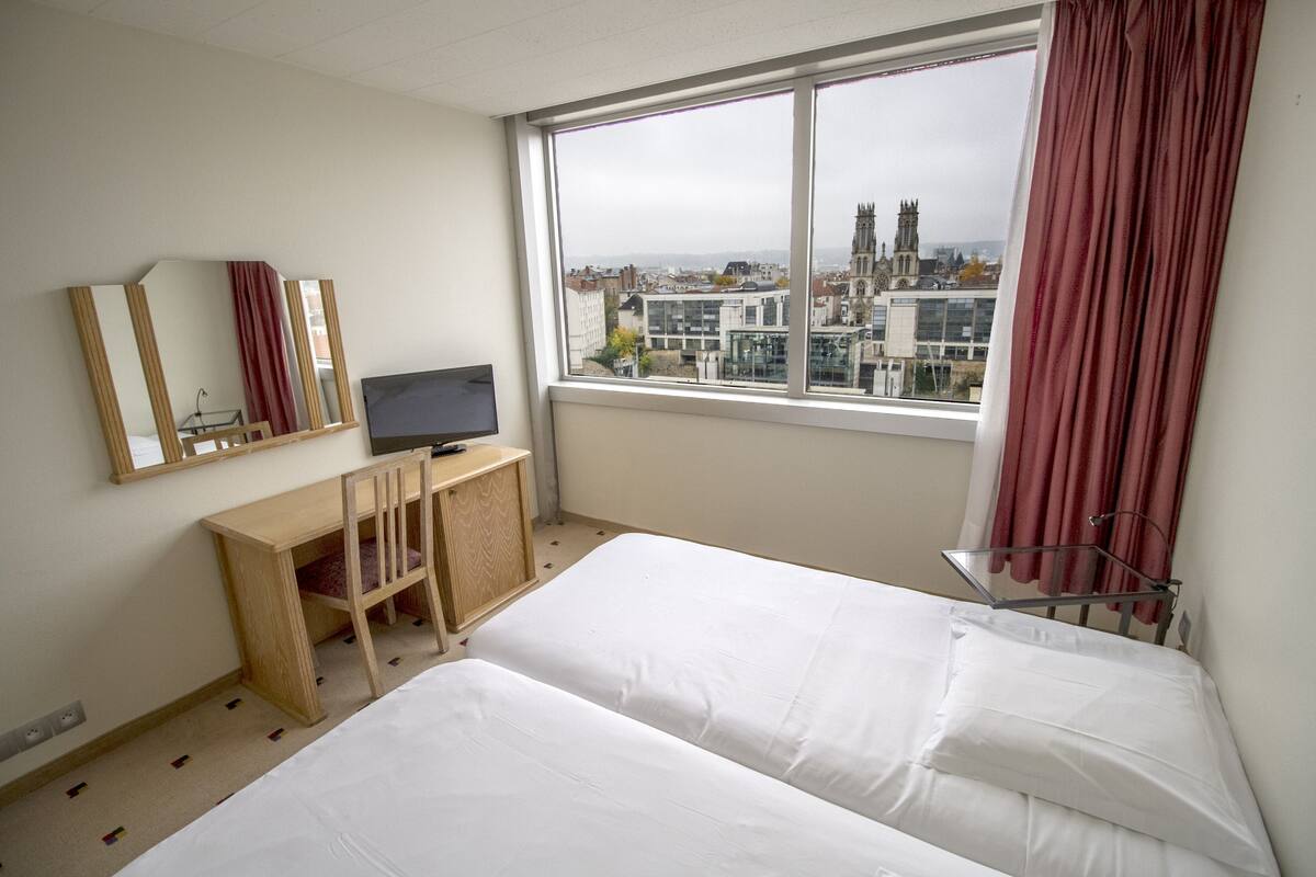 Habitación de hotel con cama, escritorio con silla, televisión y ventana con vista a edificios.