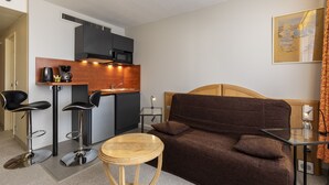 A compact hotel room with a kitchenette, a sofa, a small round table, and two bar stools. - KOSY Appart'Hôtels - Coeur de Ville  (Nancy)