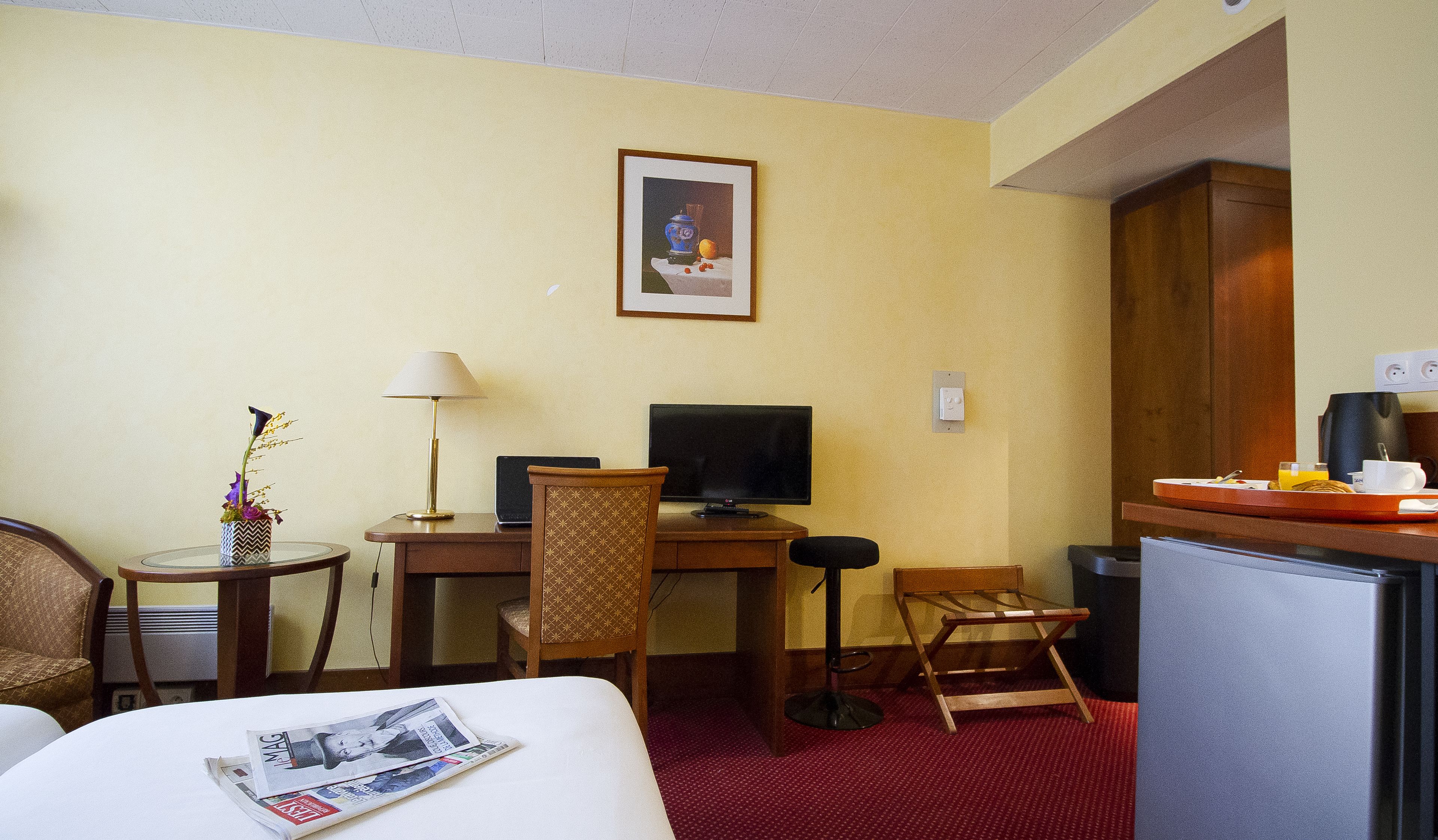 A hotel room with a bed, a desk with a computer, a chair, a small table with a lamp, a framed picture on the wall, and a kitchenette area.