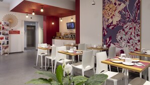 A modern dining area with white chairs and tables, a large floral mural, and a television on the wall. - KOSY Appart'Hôtels - Coeur de Ville  (Nancy)