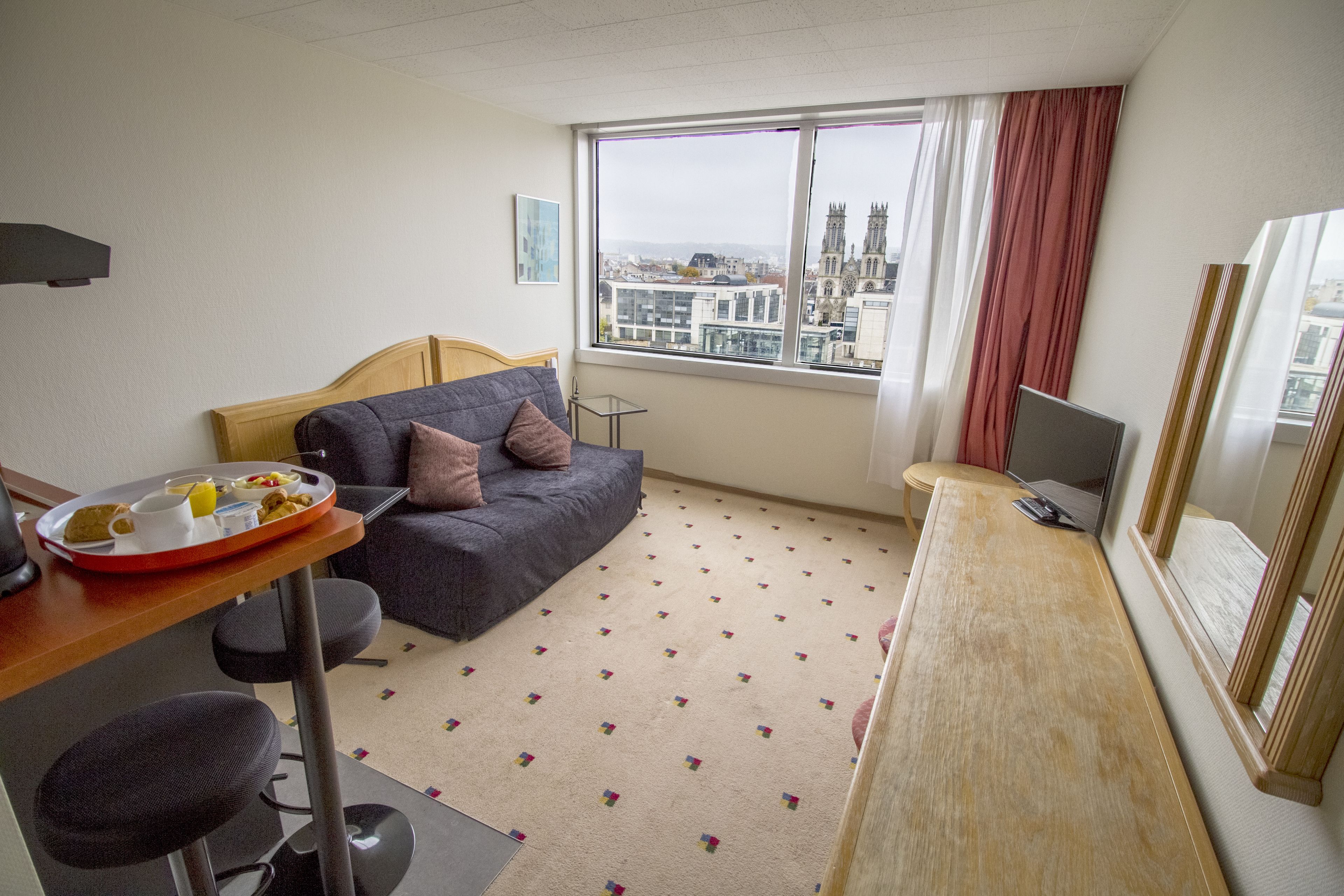 A hotel room with a bed, a desk with a computer, a dining area with a tray of food, and a large window with a city view.