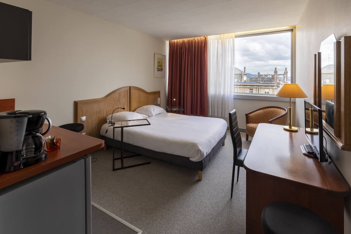 Habitación de hotel con cama, escritorio, silla, cafetera y ventana con vistas a edificios.