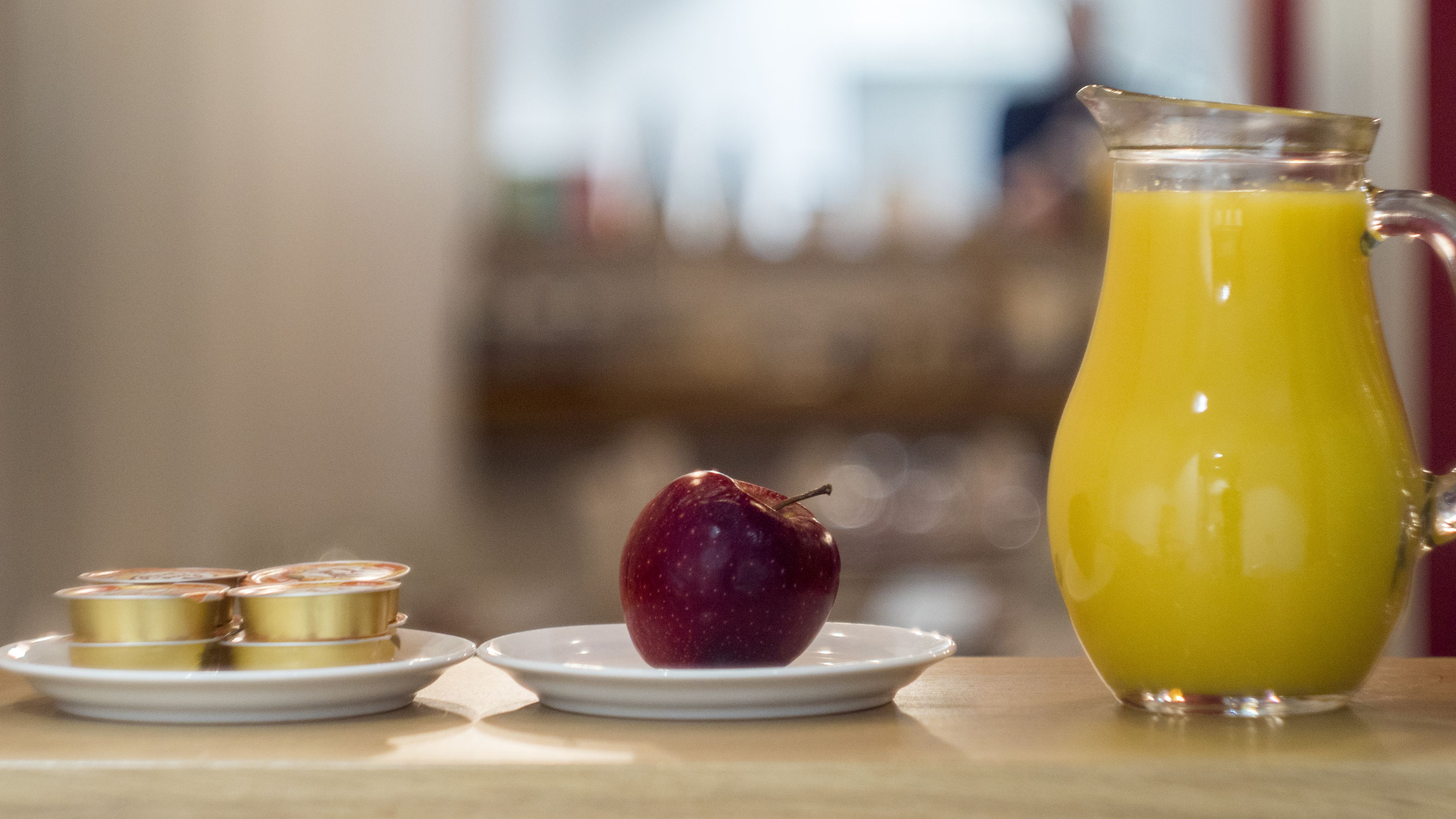 A breakfast setting with a glass pitcher of orange juice, a plate with a red apple, and two small containers on a table.