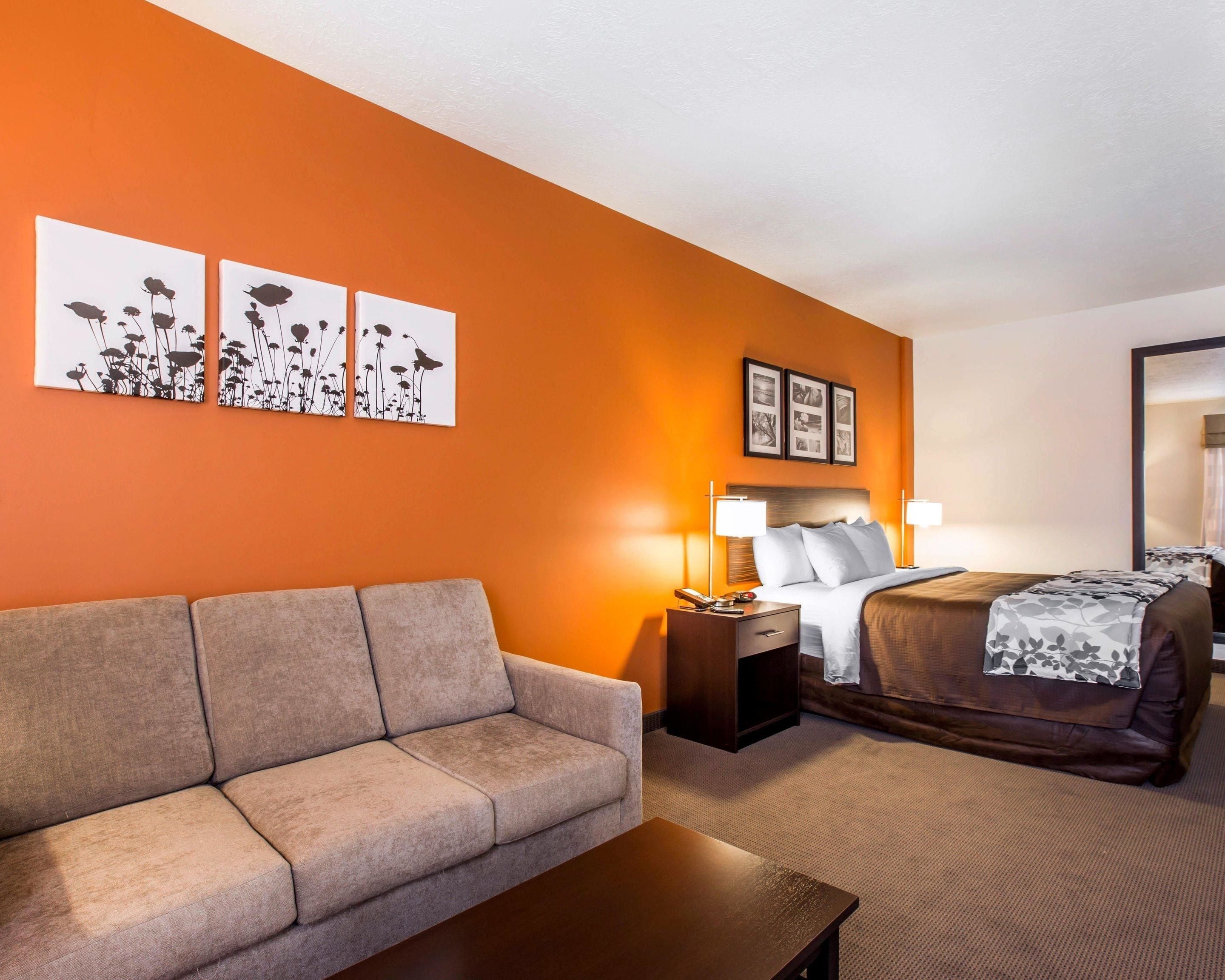 Une chambre d’hôtel avec un mur d’accent orange, un canapé, un lit recouvert d’une couverture à motifs et deux tables de chevet avec des lampes.
