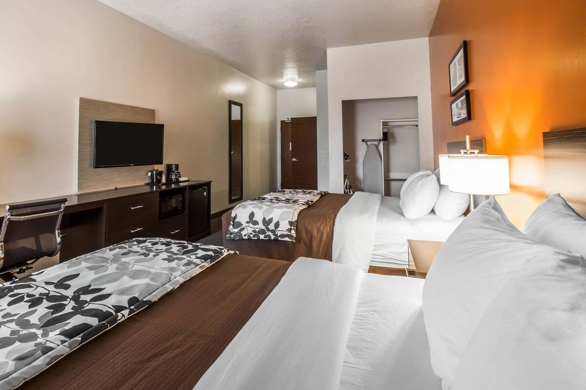 A hotel room with two beds, a desk with a flat-screen TV, a mirror, a wardrobe, and a bathroom visible through an open door.