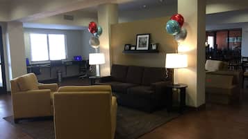 A hotel lobby with a sofa set, armchairs, a desk with a computer, and a dining area in the background.
