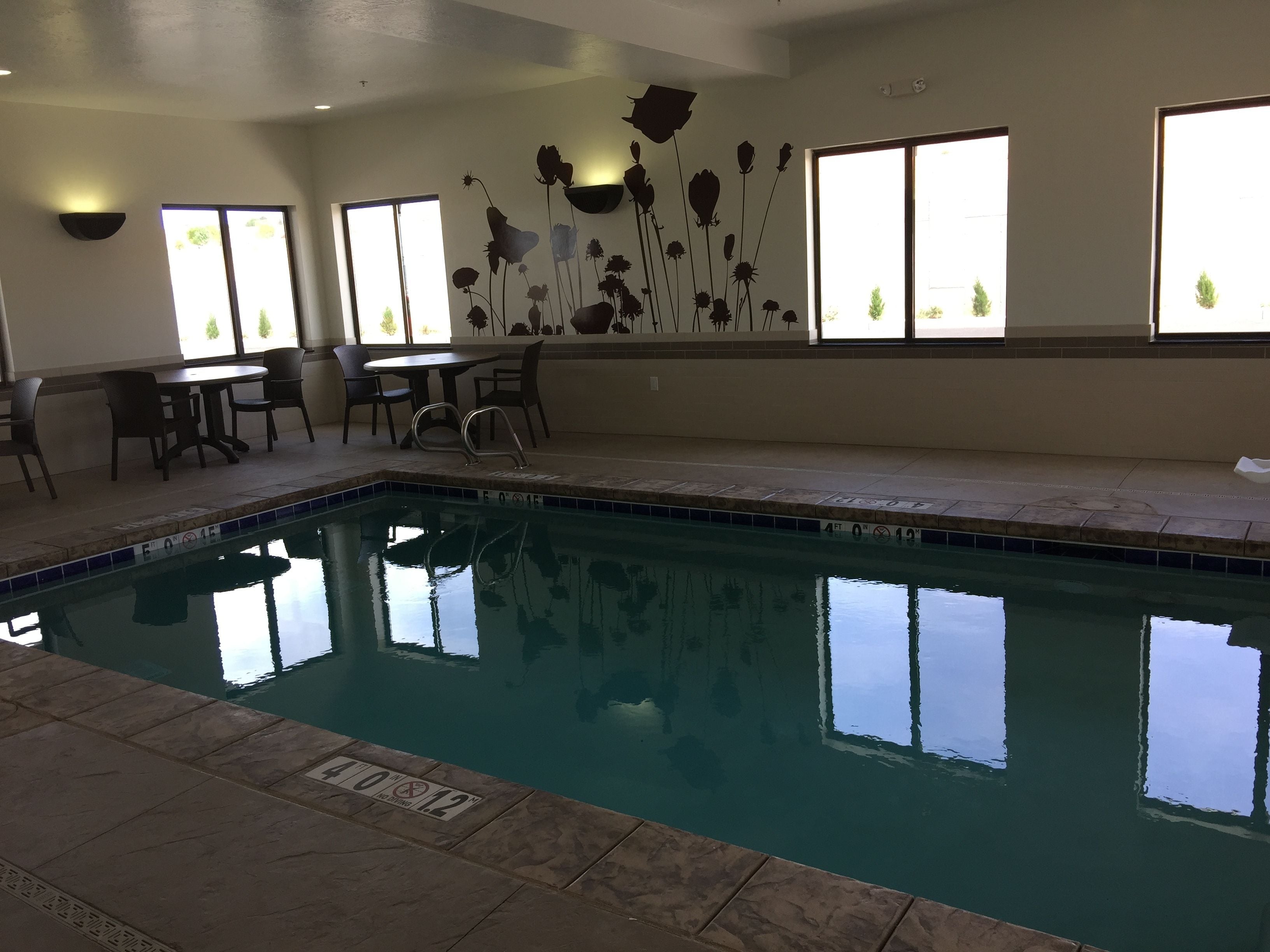 An indoor swimming pool with clear water, surrounded by a tiled deck. There are chairs and a table near the pool, and the walls are decorated with floral patterns.