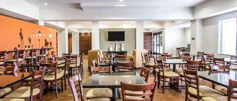 A spacious dining area with multiple tables and chairs, a flat-screen TV mounted on the wall, and a large orange mural on the left wall.