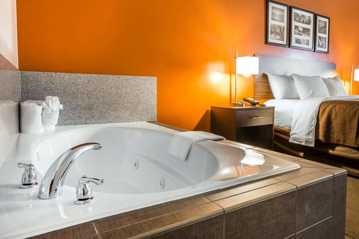A hotel room with a large bathtub, a bed with white linens, a nightstand with a lamp, and framed pictures on the wall.