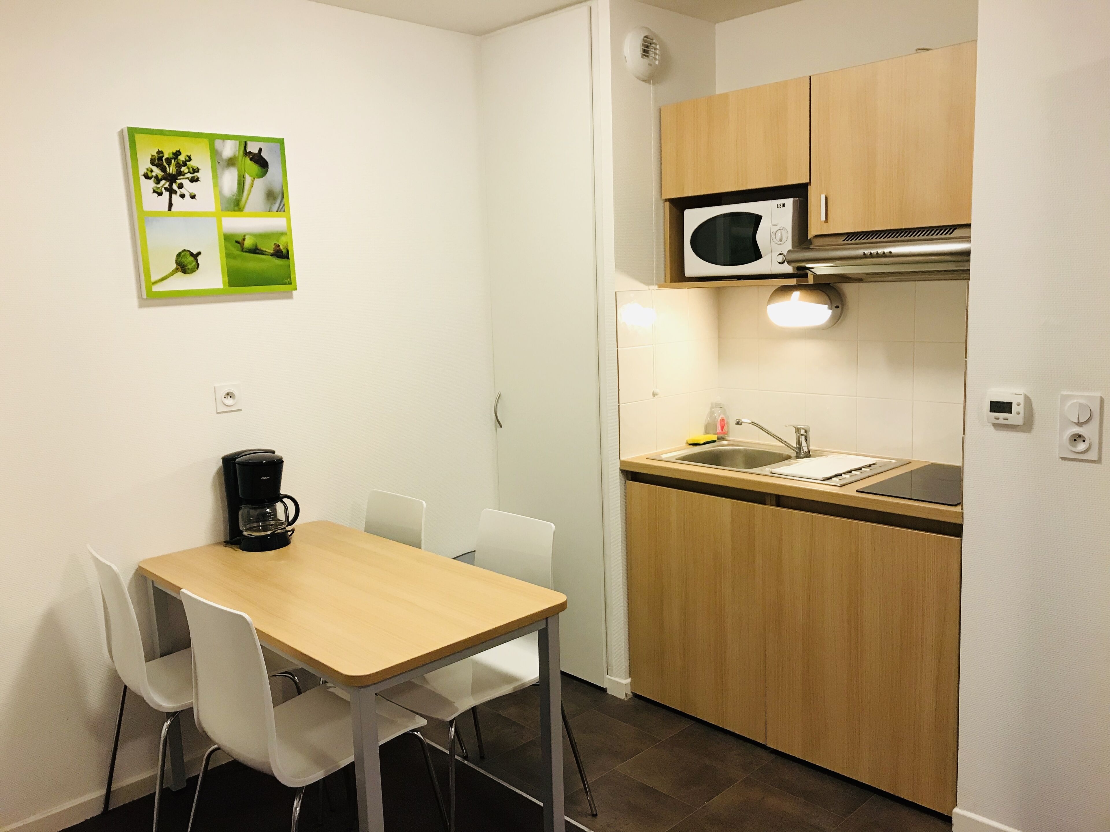 Una cocina compacta con microondas, fregadero y cafetera sobre la encimera. Hay una mesa de comedor con sillas y un cuadro colgado en la pared.