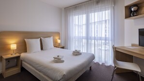 A hotel room with a large bed, two bedside tables with lamps, a desk with a chair, and a large window with sheer curtains. - KOSY Appart'Hôtels Troyes - City & Park (Troyes)