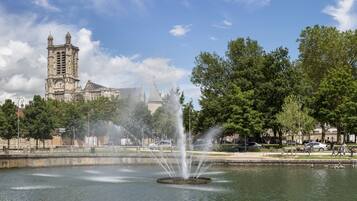 A historic building with a clock tower, a fountain in the foreground, and a park with trees and a pond.