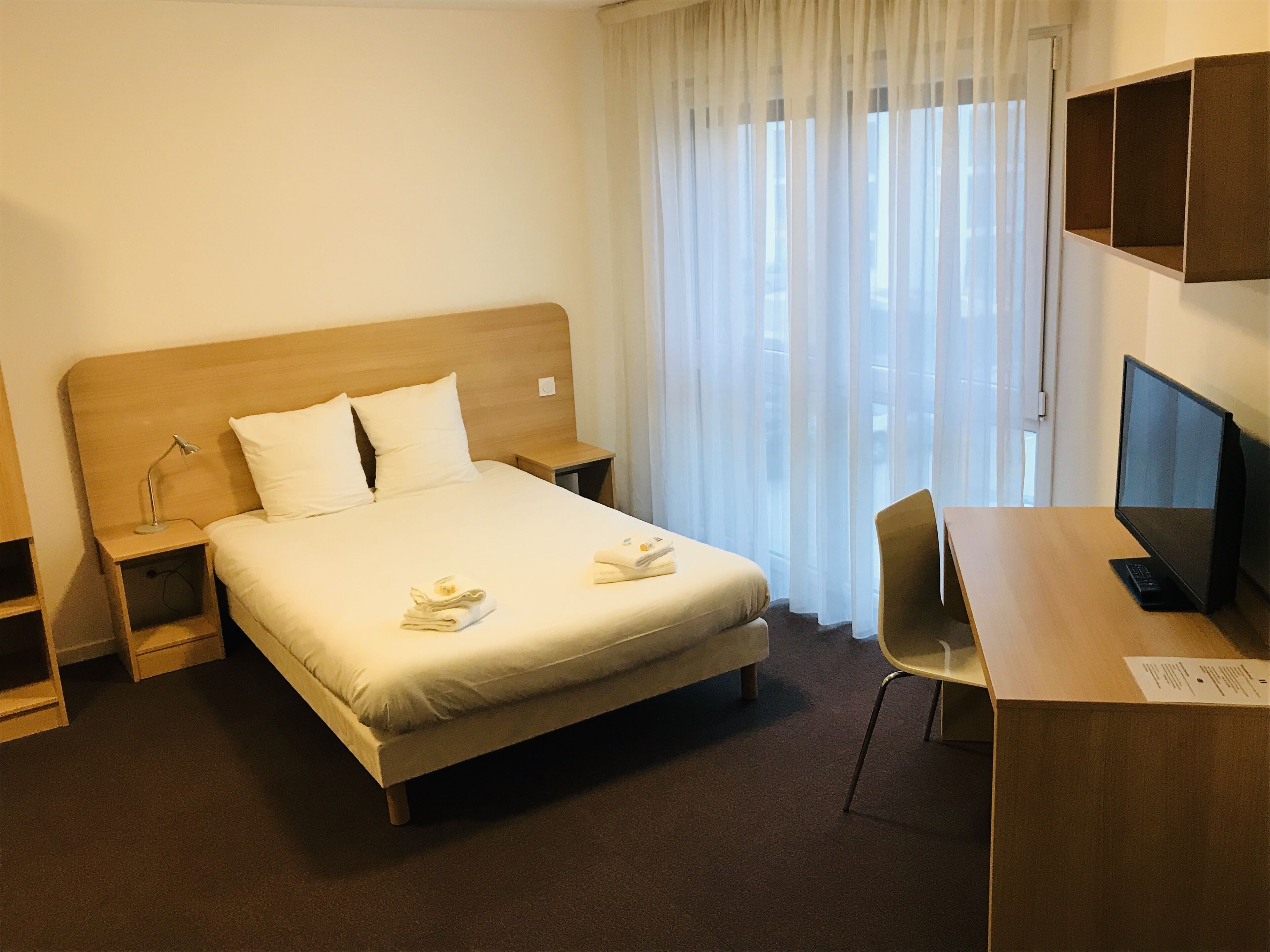 A hotel room with a bed, a desk, a chair, a TV, and a window with curtains.