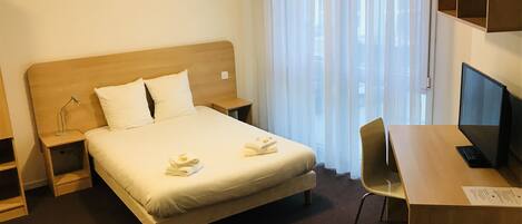 A hotel room with a bed, a desk, a chair, a TV, and a window with curtains.