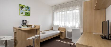 A compact hotel room with a single bed, a small desk, a chair, a TV, and a window with sheer curtains.