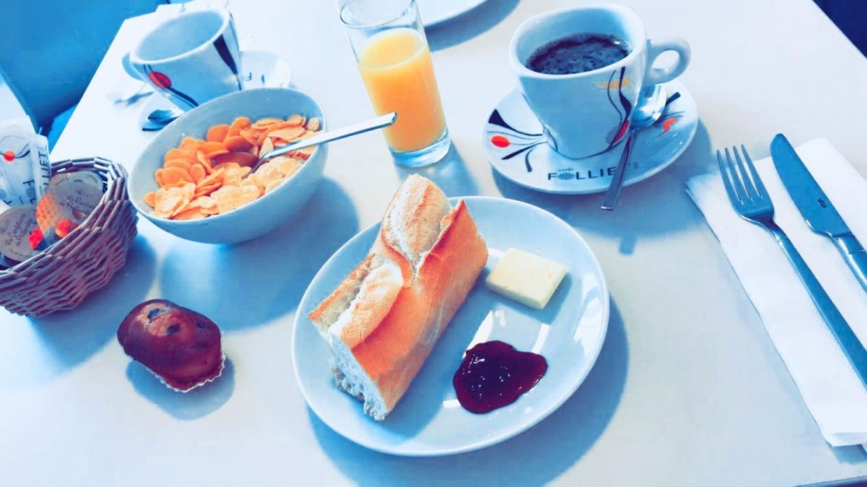Une table dressée pour le petit-déjeuner avec une assiette de croissants, du beurre, de la confiture, un bol de céréales, un verre de jus d’orange et une tasse de café.