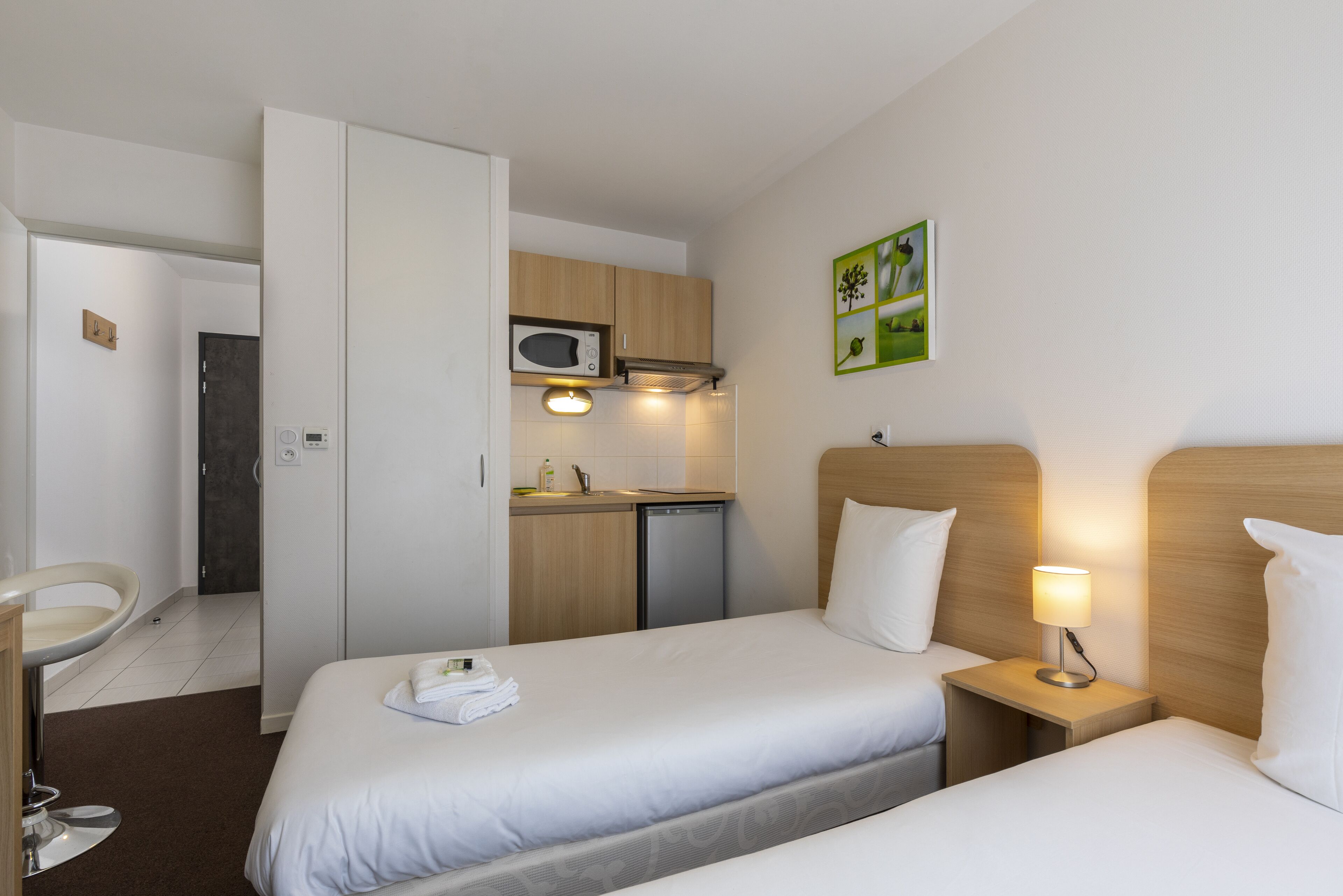 A hotel room with two beds, a small kitchenette, a desk, and a chair.