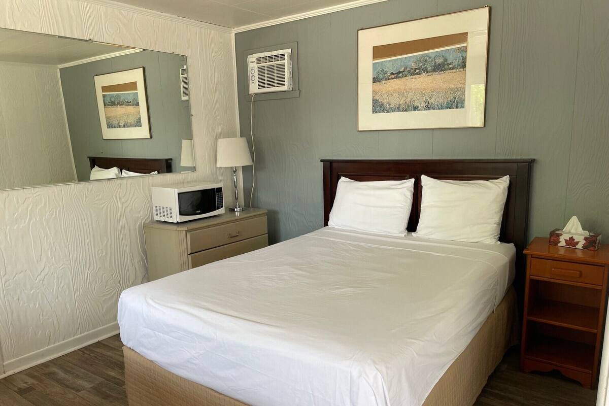 A neatly made bed with white linens, a bedside table with a lamp, a microwave, and a framed picture on the wall.