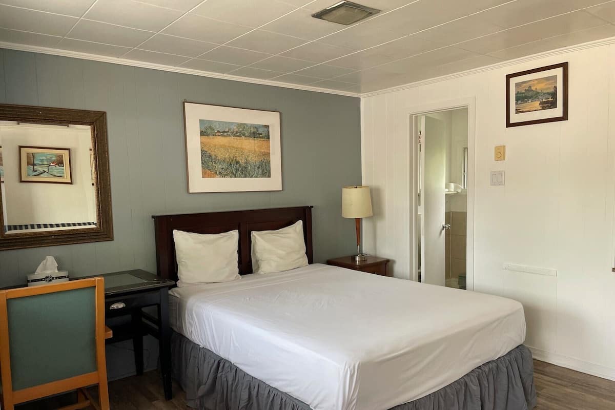 A hotel room with a bed, a desk, a chair, a lamp, and two framed pictures on the wall.