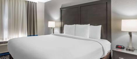 A hotel room with a large bed, two bedside lamps, a nightstand with a digital clock, and a window with curtains.