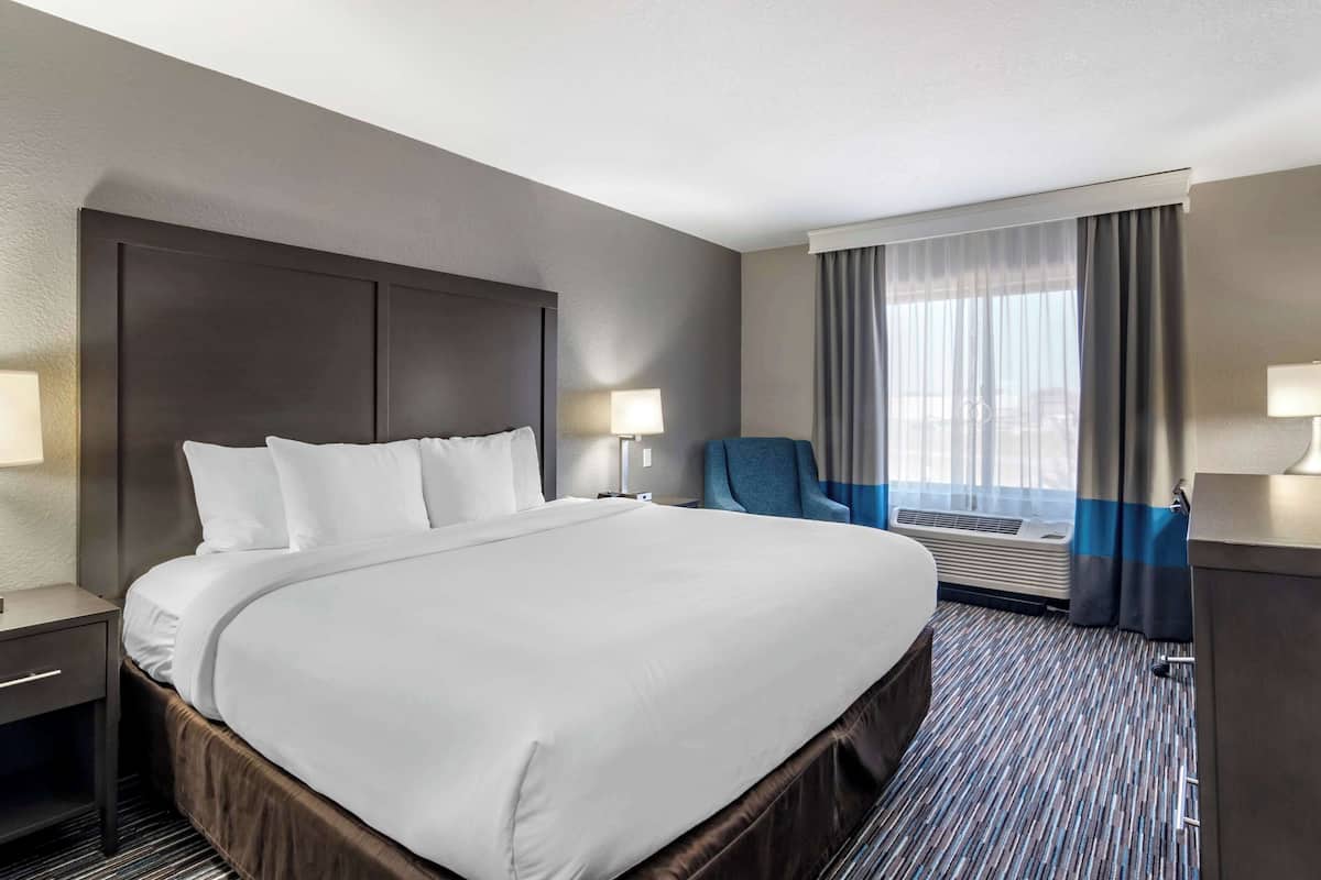 A hotel room with a large bed, a desk, and a chair. There is a window with curtains and an air conditioning unit.