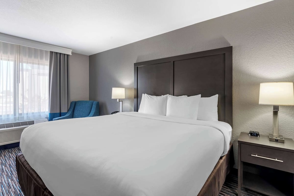 A hotel room with a large bed, two bedside tables with lamps, a chair, and a window with sheer curtains.