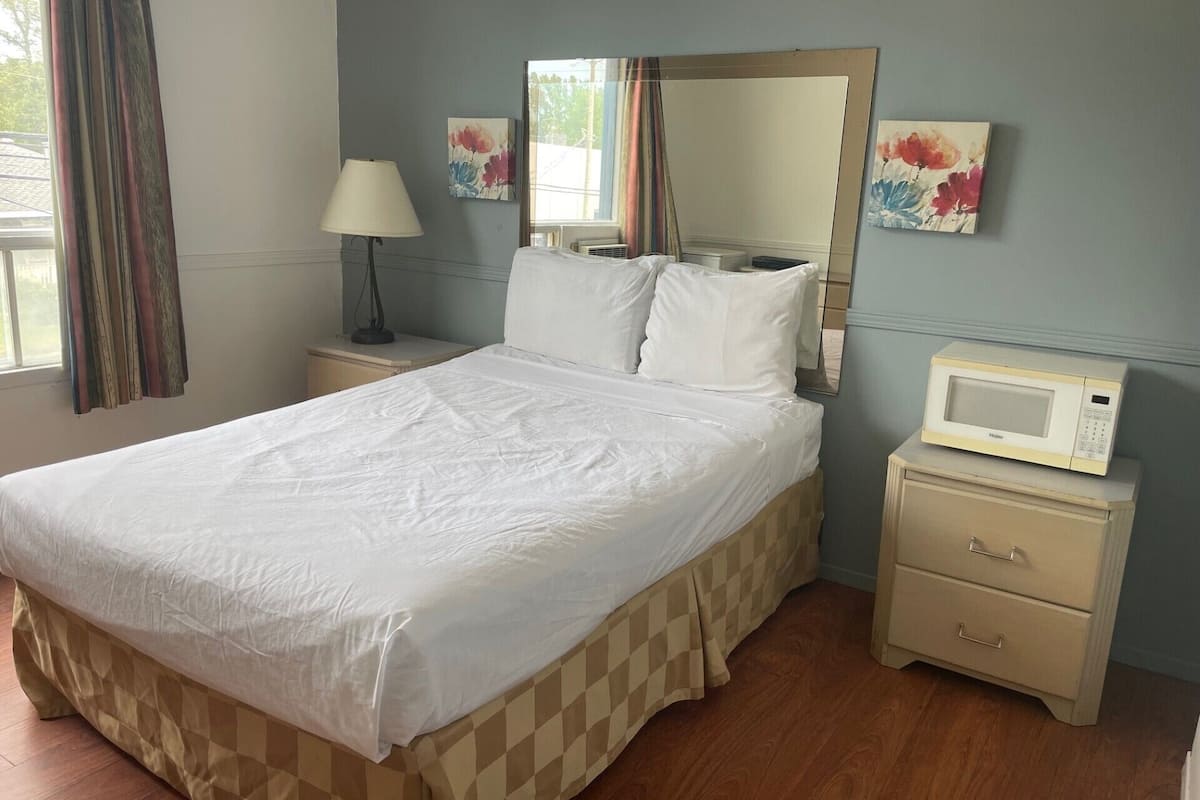 Cama bem arrumada com saia de cama xadrez, abajur, micro-ondas em uma mesinha de cabeceira e duas pinturas na parede.