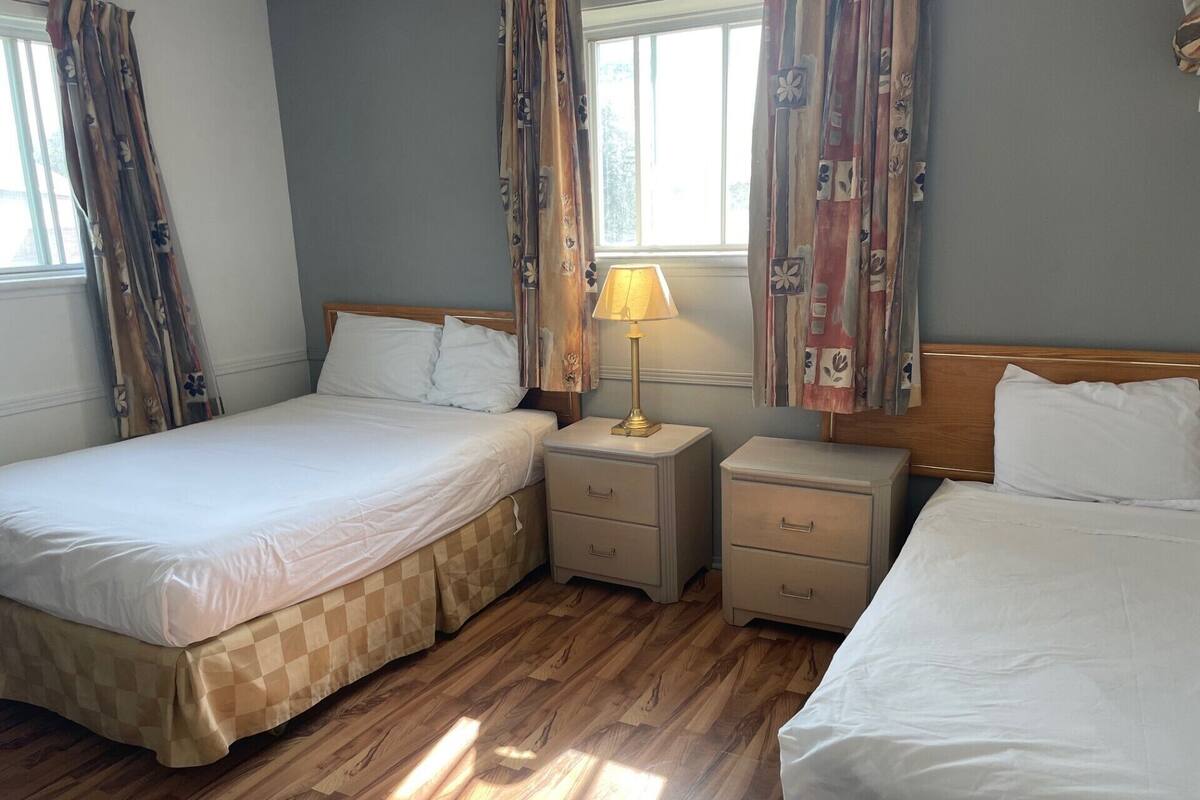 Quarto de hotel com duas camas, piso de madeira, mesa de cabeceira com abajur e cortinas.
