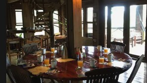 Uma mesa de jantar de madeira com cadeiras, itens de café da manhã e uma janela grande com vista.