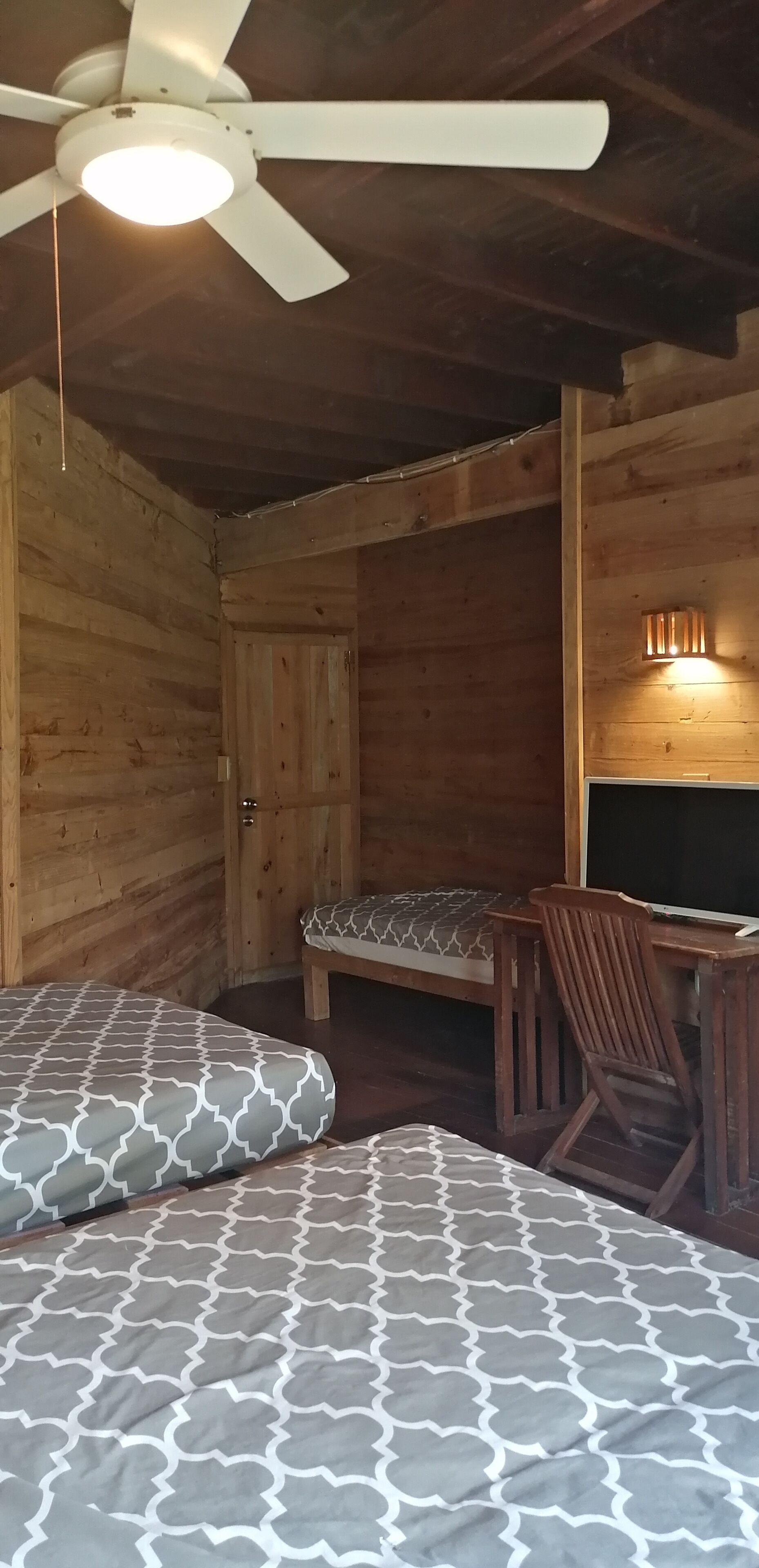 A wooden cabin room with a ceiling fan, two beds with patterned bedding, a wooden desk, and a wall-mounted light fixture.