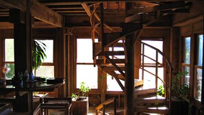 Interior rústico de madeira com escada em espiral, janelas amplas e plantas em vasos.