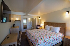A hotel room with a bed, a desk, a chair, a small table, a mirror, and a nightstand. - Harbor Inn (Santa Cruz)