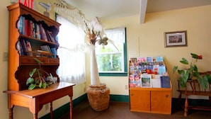 A room with a wooden bookshelf, a table with a plant, a bulletin board with magazines, and a window with curtains. - Harbor Inn (Santa Cruz)