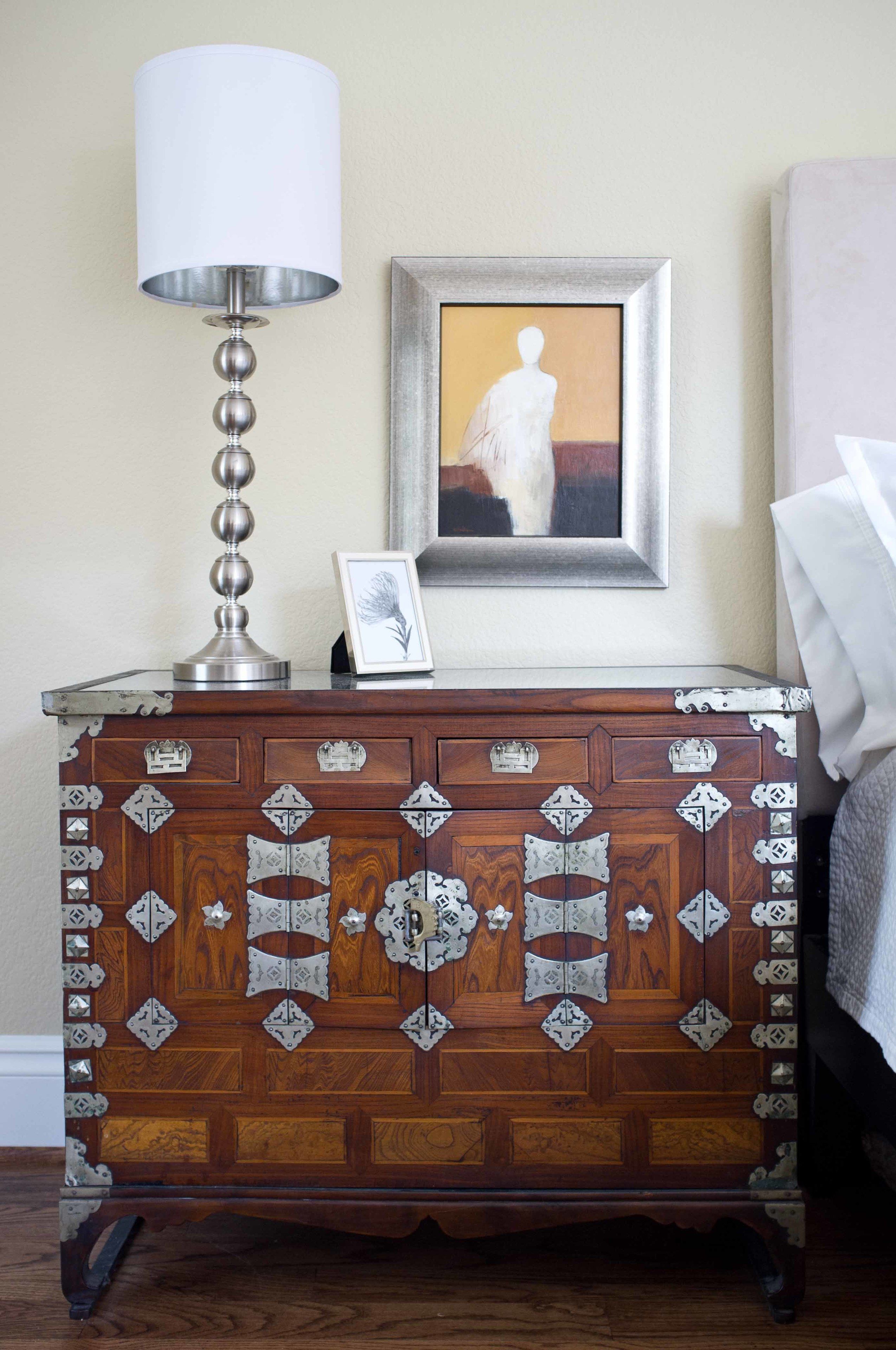 A wooden chest of drawers with metal accents, a lamp, and a framed artwork on a wall.