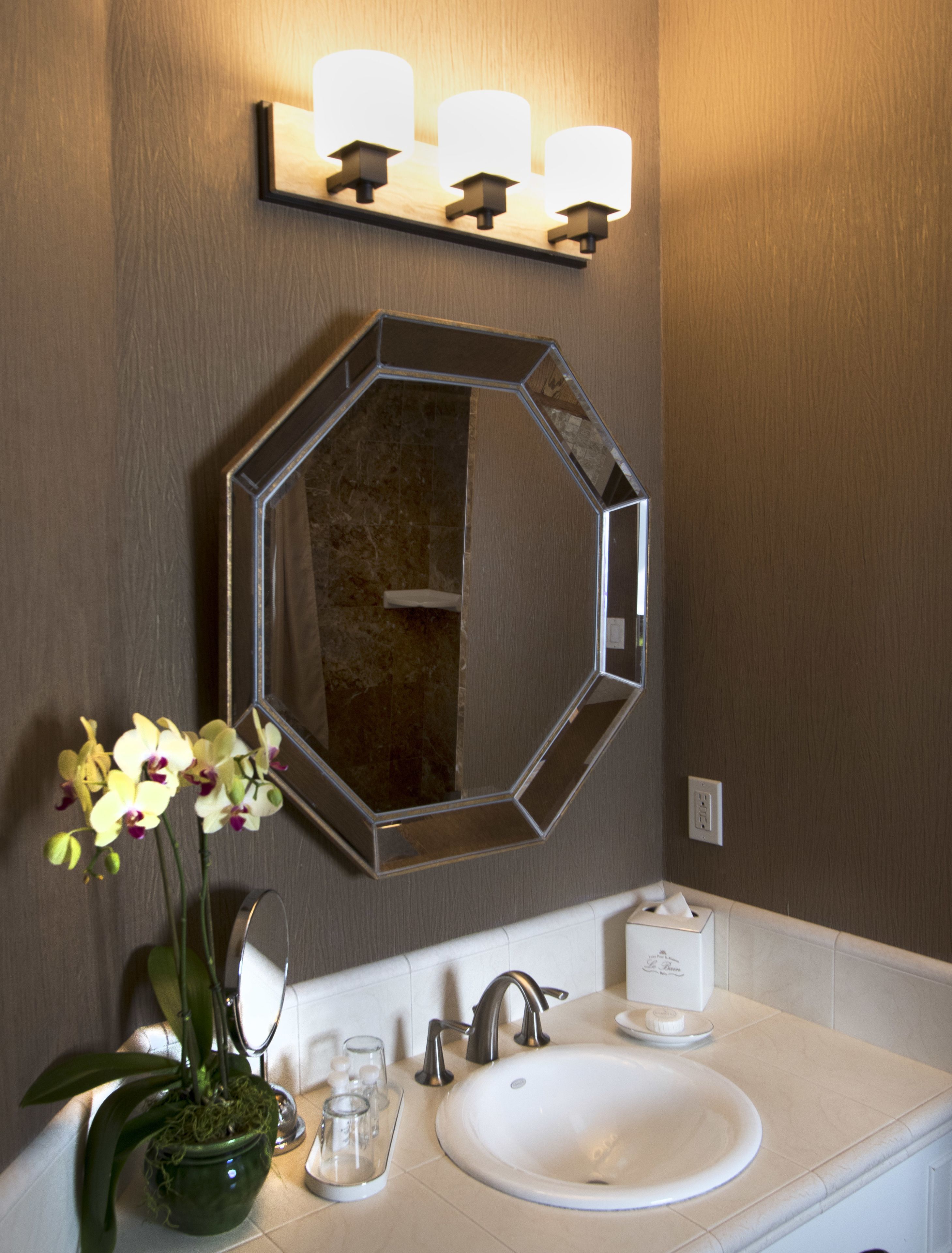 Une salle de bain avec un grand miroir octogonal, un lavabo, une orchidée en pot et un distributeur de savon.