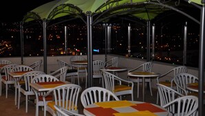 Una terraza en la azotea con mesas y sillas, cubierta por un toldo verde, con vistas a un paisaje urbano nocturno.