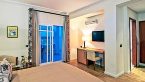 Habitación de hotel con cama, escritorio y silla, televisor y puerta corrediza de vidrio que da a un balcón.