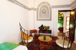 Quarto com cama, duas cadeiras, uma mesinha, um objeto decorativo na parede e um tapete estampado.