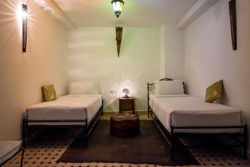 A hotel room with two single beds, a small table with a lamp, and a patterned rug.