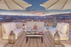 Una terraza en la azotea con dos sofás, una mesa dispuesta con comida y bebidas, y una vista de la ciudad al atardecer.