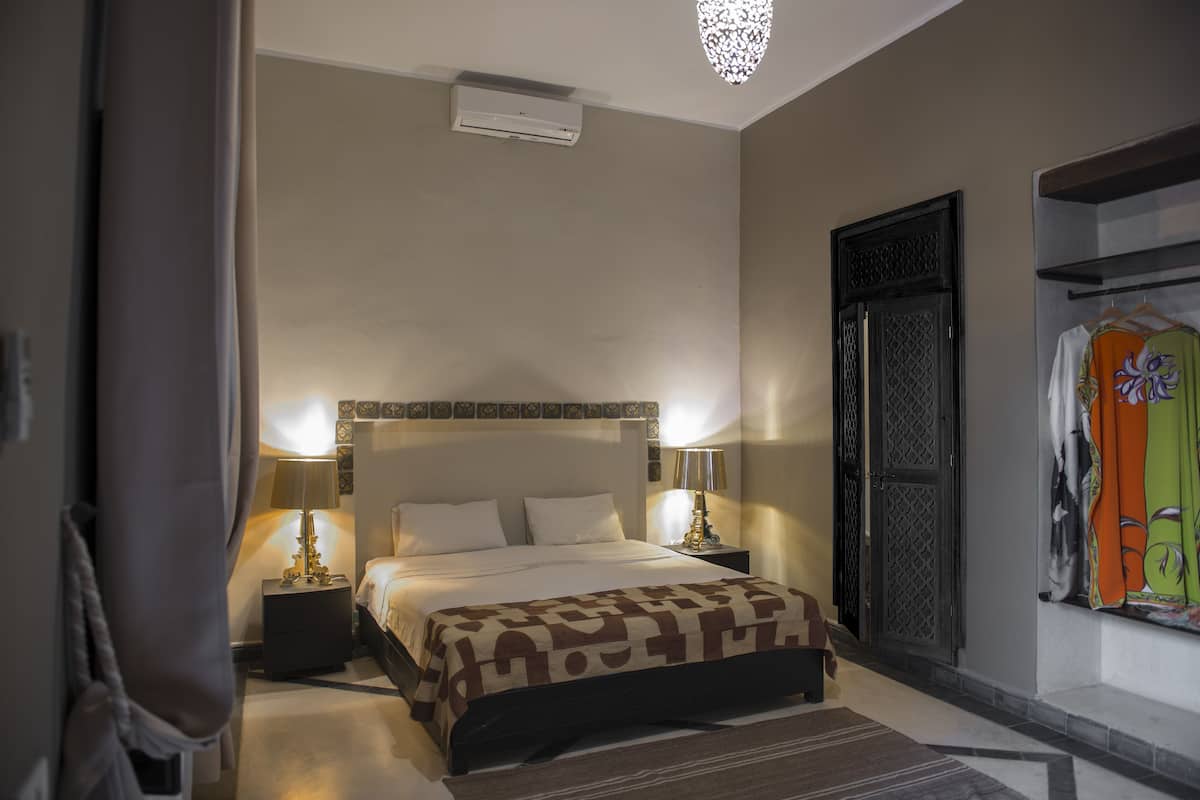 Quarto com cama, luminárias de cabeceira, ar condicionado fixado na parede e guarda-roupa com roupas coloridas.