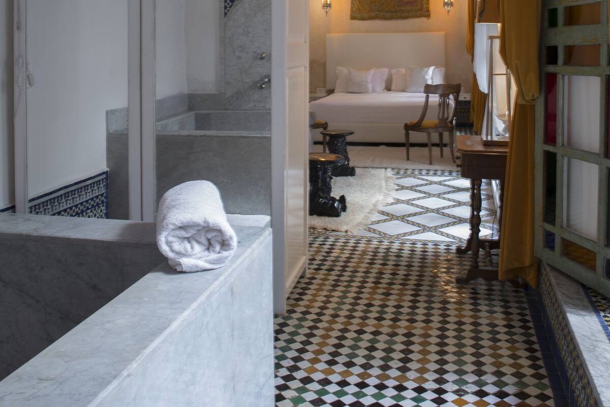 Quarto de hotel com piso em azulejo, uma cama, uma escrivaninha e banheiro com banheira e chuveiro.