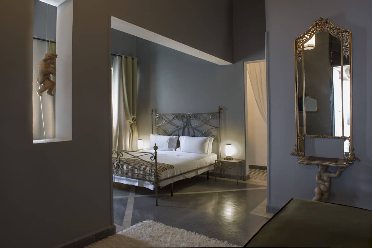 Um quarto com cama, mesas de cabeceira, um espelho e uma estátua.