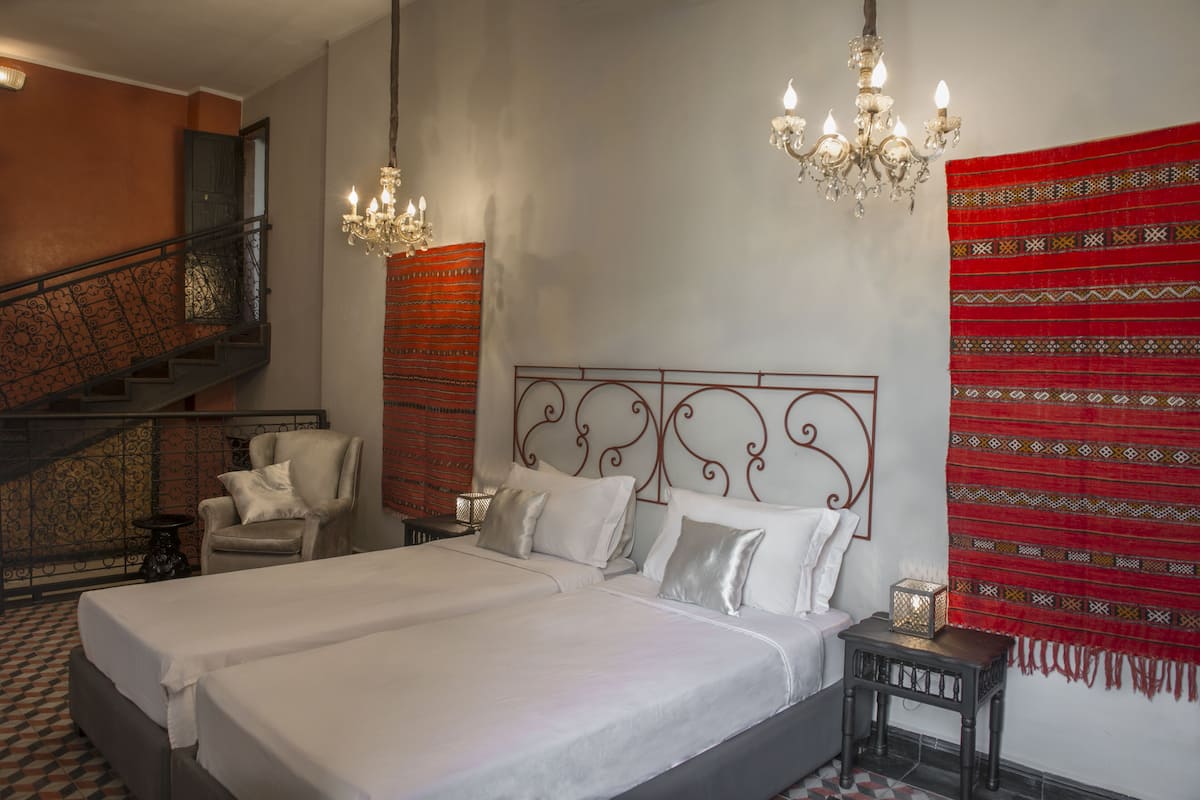 Quarto de hotel com uma cama grande, uma cadeira, uma mesinha, um criado-mudo, um lustre e uma escada.