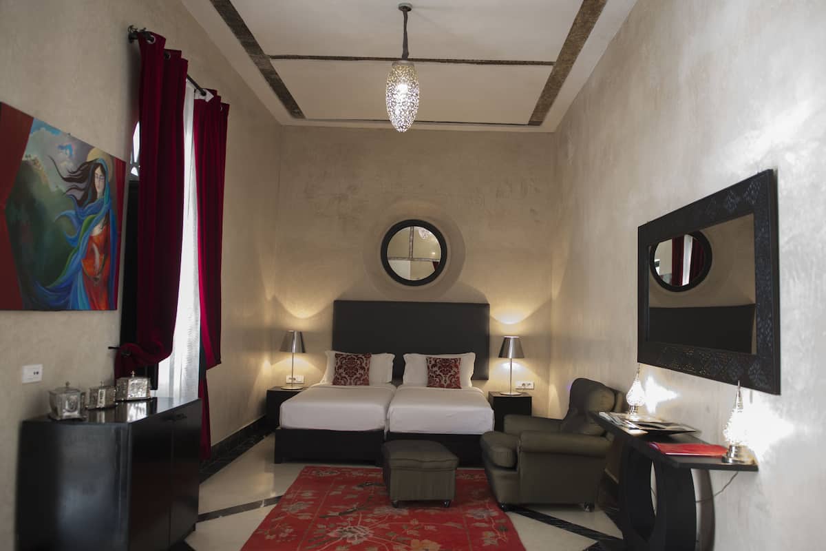 Quarto de hotel com cama, duas mesas de cabeceira, espelho redondo, tapete vermelho e um quadro na parede.