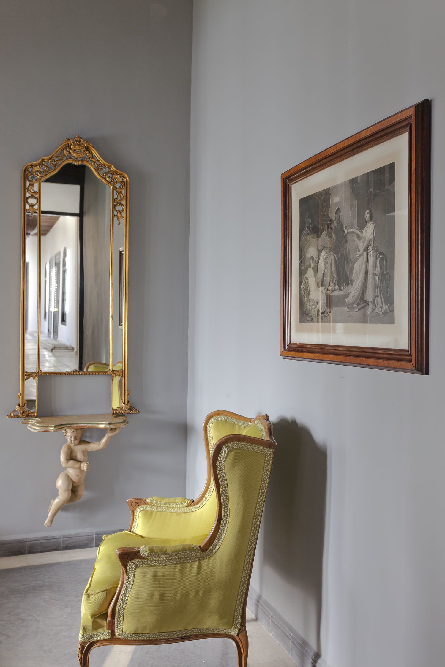 A room with a large ornate mirror, a framed black and white painting, and a vintage yellow armchair.