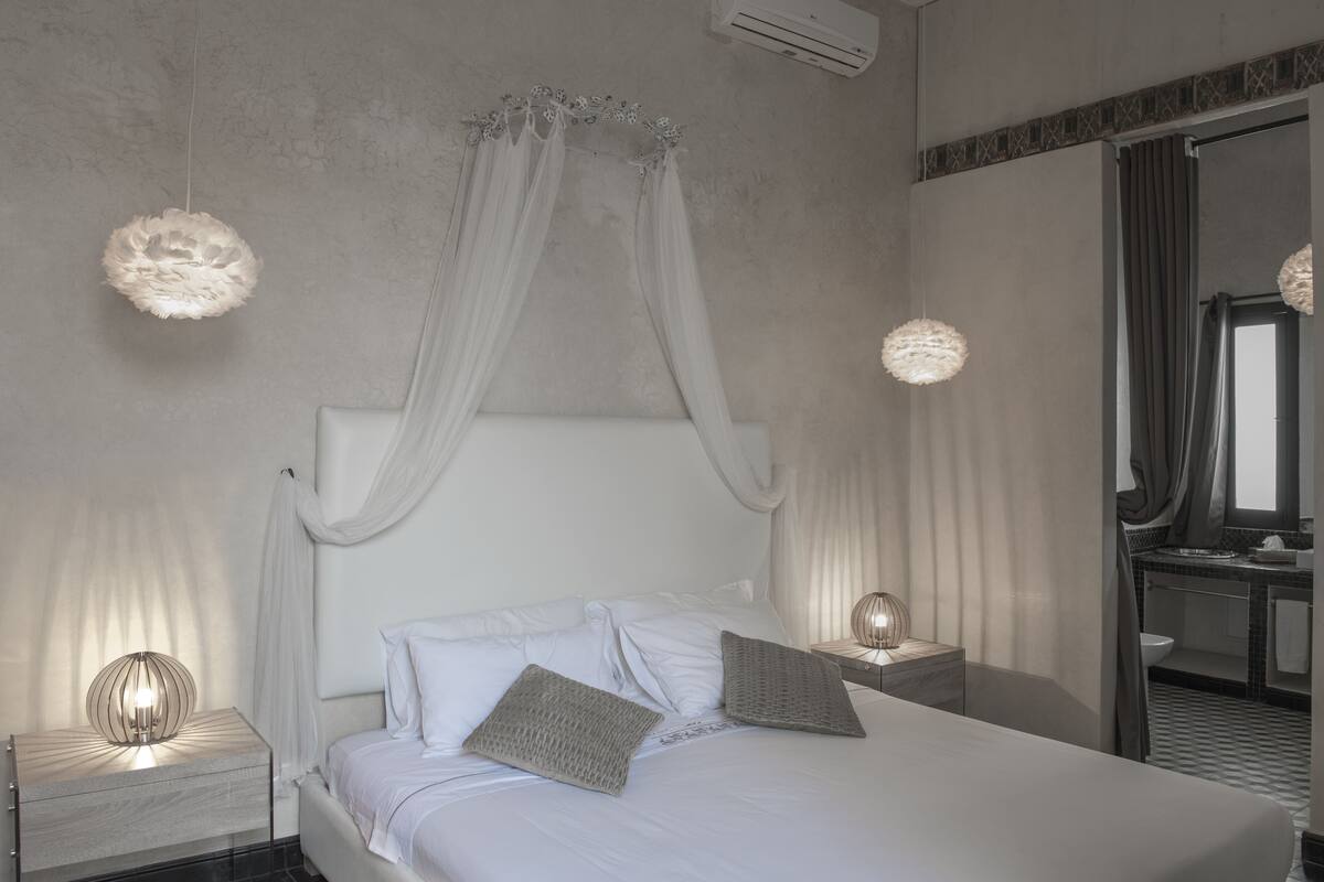 Quarto moderno com cama de dossel, abajures e banheiro visível por uma porta aberta.