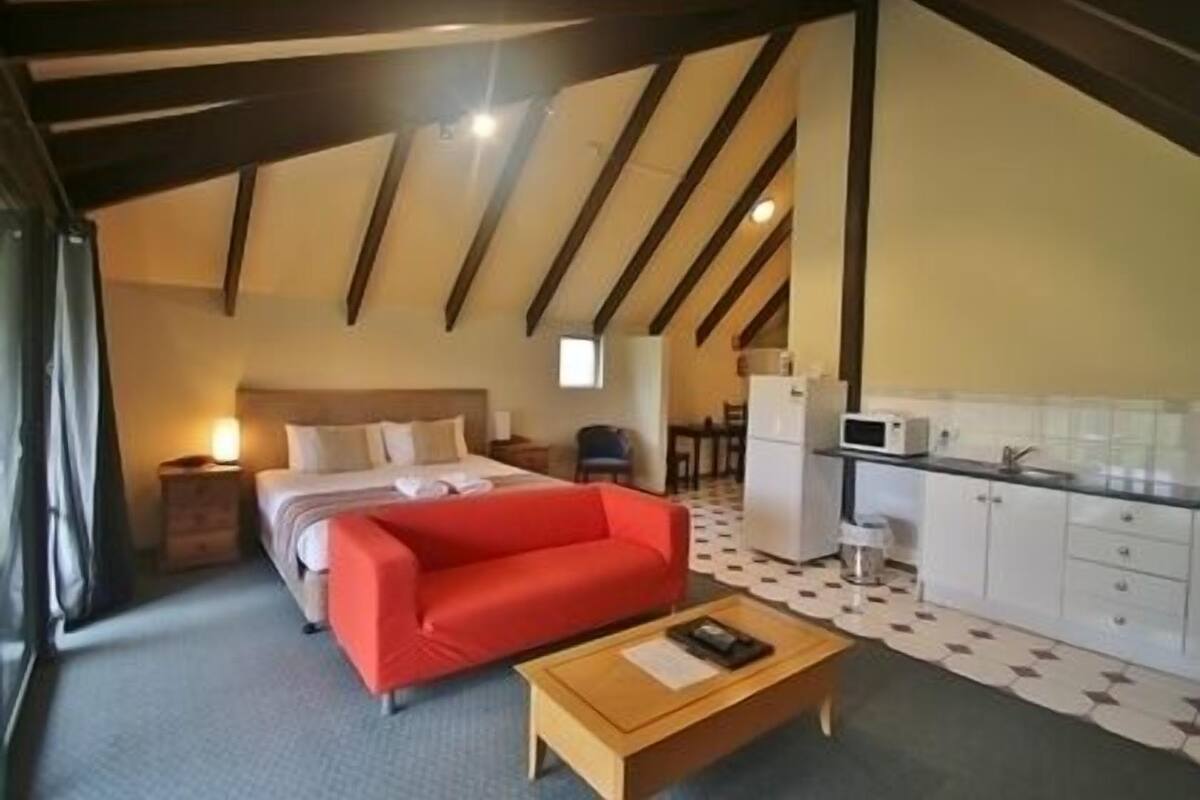 A bedroom with a bed, a red sofa, a wooden coffee table, and a kitchenette with a microwave and refrigerator.