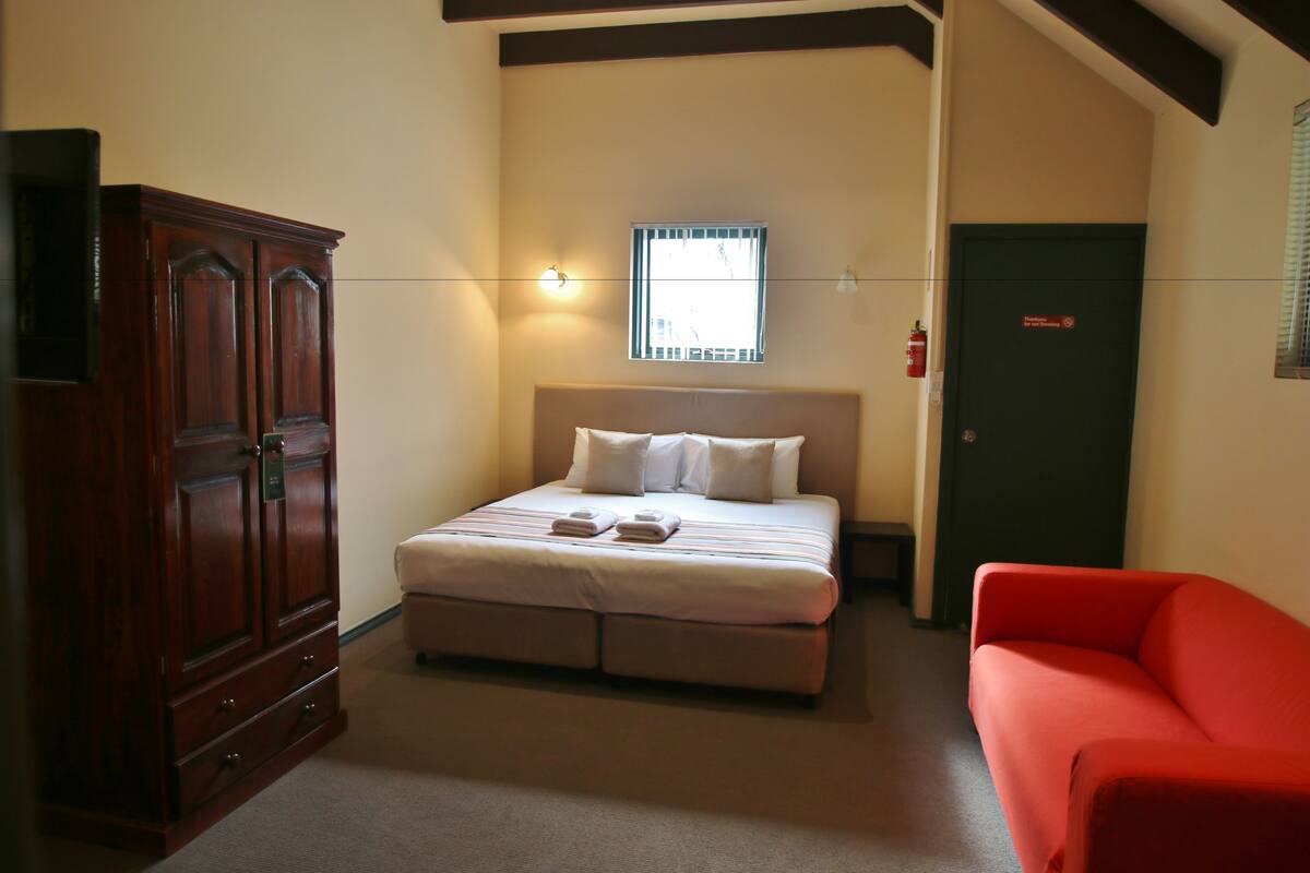 A bedroom with a bed, a wardrobe, a red chair, and a window with blinds.